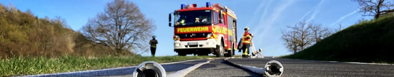 Feuerwehrmänner räumen nach einem Einsatz die Schläuche auf, im Hintergrund das Feuerwehrfahrzeug.